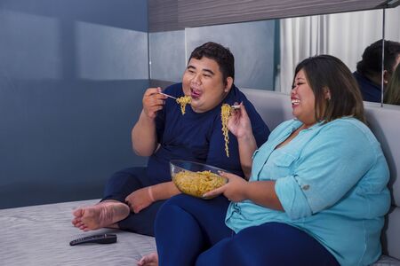 Fat Asian couple eating instant fried noodle happily, while watching television in their bedroom