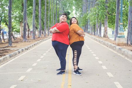 Fat Asian couple wearing sportswear while posing & looking each other by crossing their arms proudly on the road at the parkの写真素材