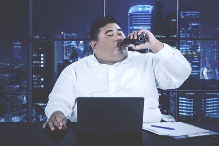 Young overweight businessman drinking cola while working overtime alone at his desk in an office at...
