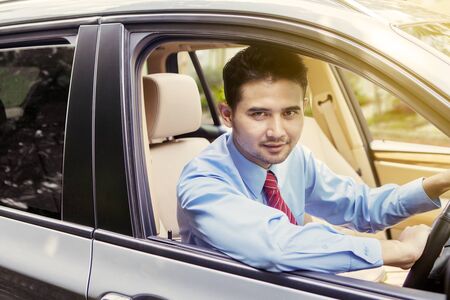 Handsome successful businessman driving a car while looking and smiling at the camera through the windowの写真素材