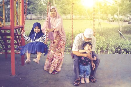 Elderly Asian muslim grandparents playing with their grandchildren, while swinging in the playground...