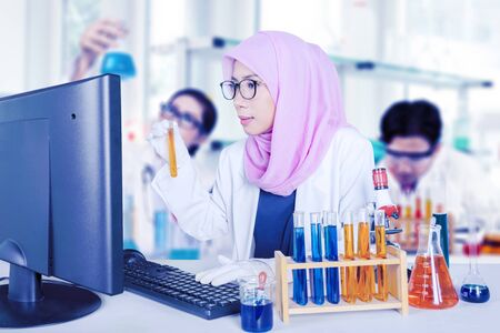 Muslim scientist doing experiment with chemical tube while looking at computerの写真素材
