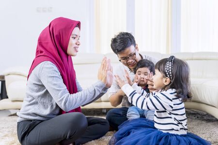 Happy Asian muslim family sitting on a rug while clapping hands together playfully in their living...