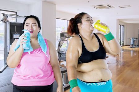 Two overweight women drinking water while standing in the fitness center after exercisingの写真素材