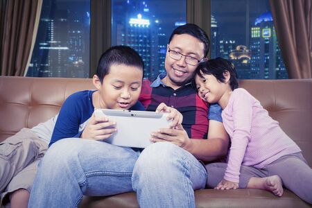 Portrait of handsome Asian man sitting on a couch while teaching his children how to use a tablet...