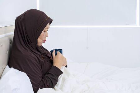Side view of sick old woman holding a glass of tea while resting on the bedの写真素材