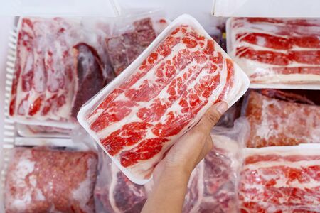 Top closeup of human hand picking a package of big chunk red meat, plastic wrapped from the freezer box in the storage roomの写真素材