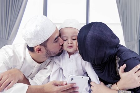 Young muslim parents kissing their son while using a mobile phone on the bed at home