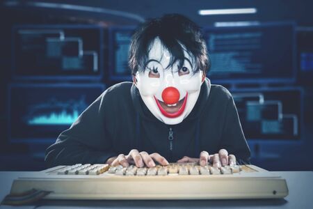 Male hacker working with computer while wearing a clown mask