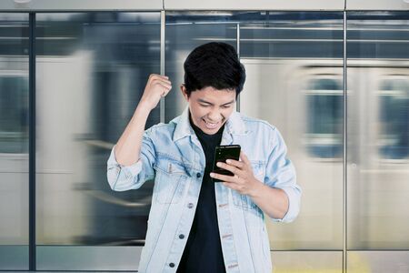 Excited young man reading good news on cellphone while standing in the train station