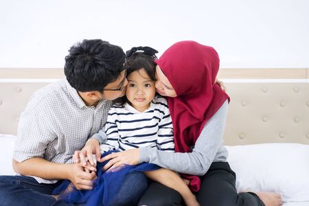 Portrait of young muslim parents kissing their cute daughter while sitting on the bedの写真素材