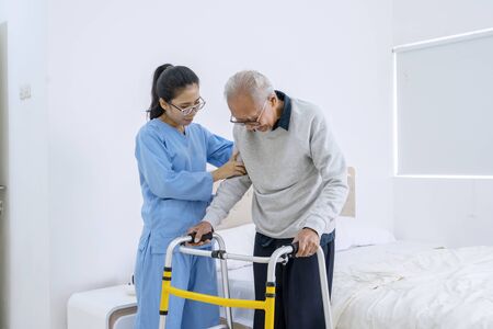 Portrait of beautiful nurse helping old man to walk using walker equipment in the bedroomの写真素材