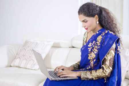 Portrait of beautiful indian woman wearing saree clothes while using laptop in living roomの写真素材