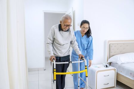 Picture of young nurse helping old man to walk with walker equipment while standing in the bedroomの写真素材