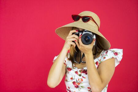 Portrait of fashionable tourist taking photo using digital camera while wearing hat in studio with red background の写真素材