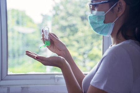 Close up of young woman applying hand sanitizer while wearing medical mask at homeの写真素材