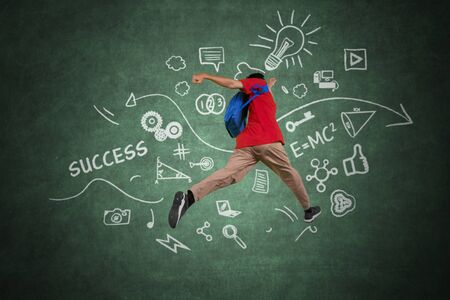 Picture of male college student leaping with doodles on the blackboard while carrying rucksack 