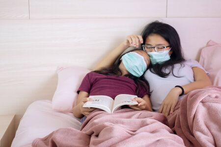 Portrait of little daughter and her mother sleeping on the bed while wearing face mask and holding a bookの写真素材