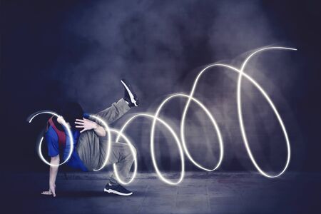 Picture of cool boy doing break dance while wearing hat and carrying backpack with light background