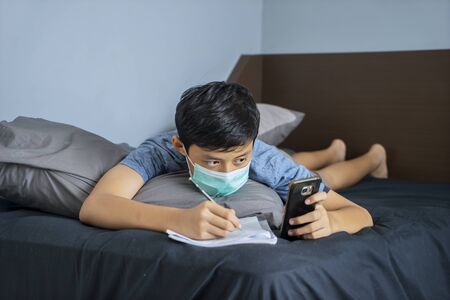 Portrait of elementary student studying at home using cellphone while wearing medical maskの写真素材