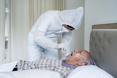 Portrait of unidentified doctor wearing hazmat suit and face mask while checking up his old Asian patient's mouth on the bed in the patient's roomの写真素材