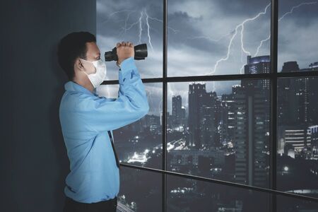 Side portrait of unidentified man wearing a blue suit and face mask, while peeking at the thunderstorm outside with his binoculars in his officeの写真素材