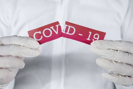 Closeup of unknown hands wearing gloves while holding a torn red label written "Covid-19"の写真素材