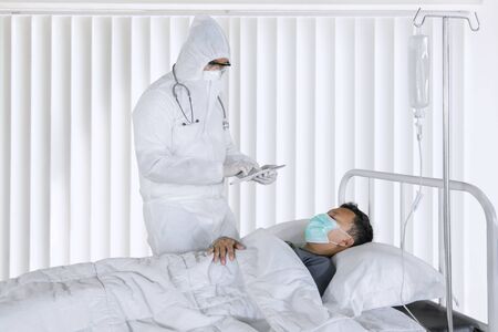 Portrait of male doctor examining young patient using digital tablet while wearing protective suit in hospitalの写真素材