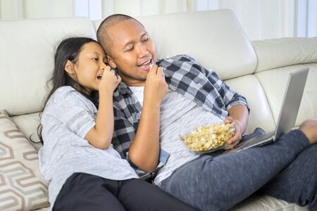 Little girl and her father watching a movie on a laptop computer while enjoying popcorn on the couch. Shot at homeの写真素材