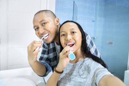 Young man and his daughter taking selfie picture together while brushing teeth in the bathroom. Shot at homeの写真素材