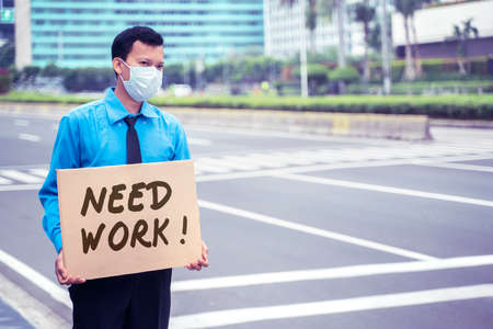 JAKARTA - Indonesia. May 12, 2020: Male employee wearing a medical mask while holding text of need work on a cardboard and standing on the sidewalk. Covid-19 effect conceptのeditorial素材