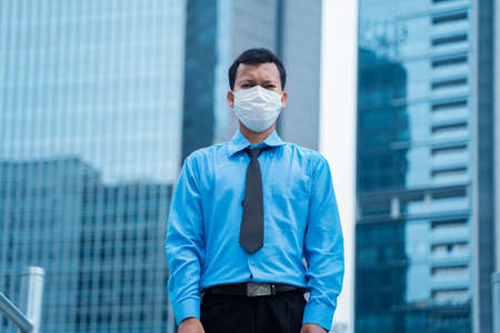 JAKARTA - Indonesia. May 12, 2020: Male employee wearing a mask to prevention coronavirus while looking at the camera and standing with skyscrapers backgroundのeditorial素材