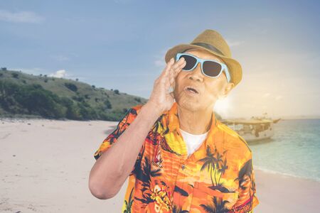 Old man wearing sunglasses and summer clothes while shouting on the beach with sunlight backgroundの写真素材