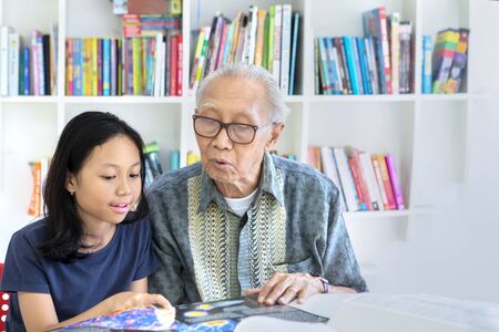 Senior man helping his granddaughter to study while studying together in the library with bookcase backgroundの写真素材