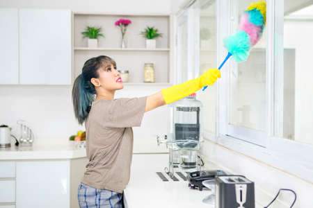 Beautiful woman is cleaning windows with a duster brush and standing in the kitchen. Shot at home