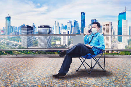 Young businessman wearing a mask and sitting in the chair while working with cellphone and laptop at office rooftopの写真素材