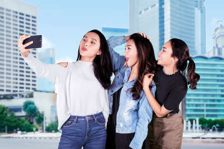 Group of teenage girls using a smartphone to take a selfie photo together on the roadside with skyscrapers backgroundの写真素材