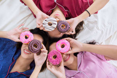 Top down view of three teenage girls having sleepover party while making funny face with donuts and lying on the bed at homeの写真素材