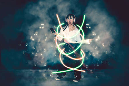 Young woman listening music on mobile phone using headphone while dancing with neon light and smoke...