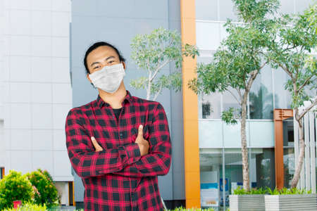Male college student looks confident with folded arms while wearing a mask and standing at university yardの写真素材