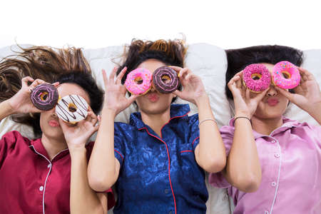 Top down view of three teenage girls covering their eyes with donuts on the bed while having...