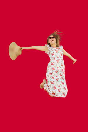 Excited Caucasian woman jumping in the studio while wearing sunglasses and hat over red background