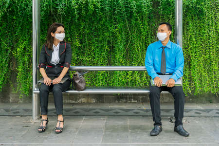 Young business couple wearing mask and chatting together while sitting separately on bus station during new normal lifestyleの写真素材
