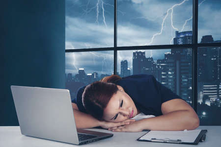 Fat businesswoman sleeping in the office while working with a computer and paperwork on the desk with stormy city backgroundの写真素材