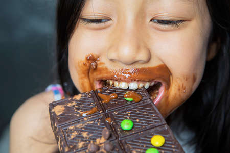 Little girl looks happy while biting and eating a bar of chocolate with messy her mouth
