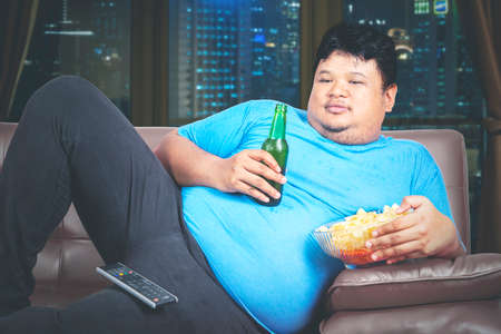 Obesity man eating chips and drink a beer in the living room while watching TV and lying on the sofa. Shot in the apartmentの写真素材