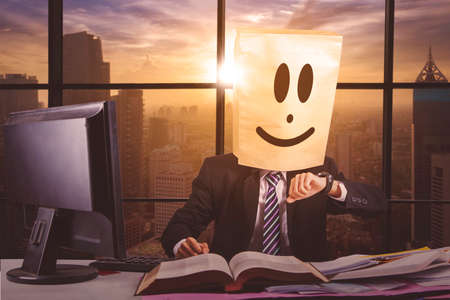 Overworked businessman working in office with cardboard head and looking at his watch with sunset...