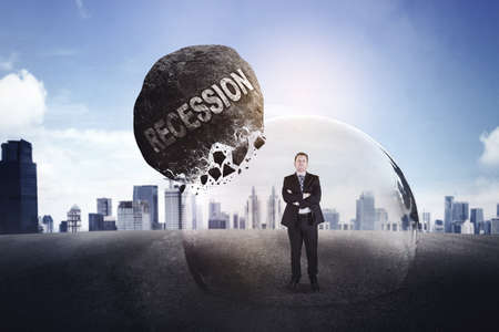 Confident businessman looking at the camera while standing in bubble to protect from falling stone with recession word. Shot with modern city backgroundの写真素材