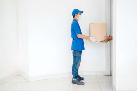 Side view of delivery man wearing face mask and gloves while carrying a stack of packages and standing near an opened door of customer houseの写真素材