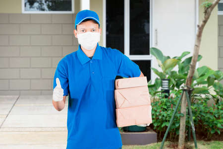 Side view of delivery man wearing face mask and gloves while carrying a stack of packages and standing near an opened door of customer houseの写真素材